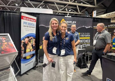 Two women in navy shirts smile and pose at a trade show booth promoting GearWash PPE Rentals for first responders with banners in the background.