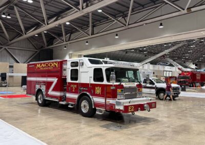 Fire engine on display in a large indoor exhibition hall with metal trusses and bright ceiling lights.