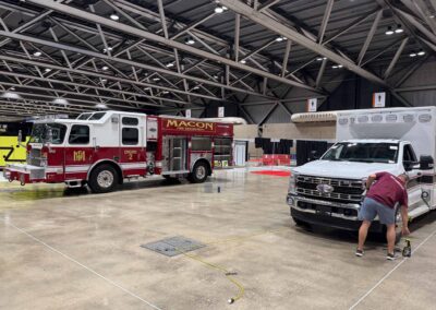 Indoor exhibition hall with multiple fire trucks, including a red Macon Fire Department engine, and a person kneeling near a white truck.