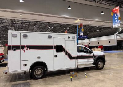 White ambulance with a red stripe parked inside a large exhibition hall under hanging lights and banners.