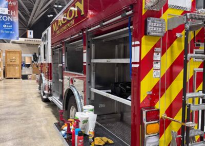 Red fire truck on display at an indoor expo, with open side compartments and gear on the rear step.