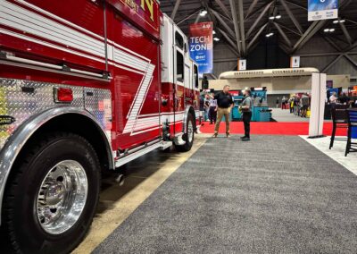 Red fire engine on display at an indoor expo, with people inspecting it near a red carpet and banners in the background.