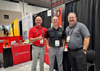 Three men standing at a Streamlight booth, smiling for a photo among red-draped tables and product displays at a trade show.