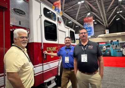 Three men pose beside a red fire engine at a convention, with tech zone banners in the background.