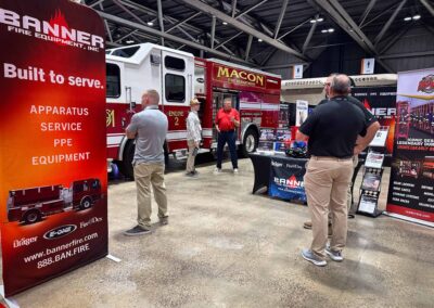 Trade show booth featuring a red Banner Fire Equipment banner beside a white and red fire truck; several men stand and converse in a convention hall.