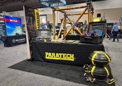 Paratech exhibit at a trade show with yellow steel lifting rig and gear cases, attendees conversing in the background.