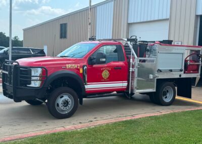 Red fire department brush truck with 'Brush 2' on the door, parked by a beige industrial building with equipment on the back.