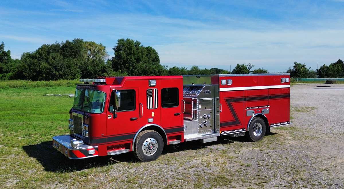 Red fire engine parked on a grassy field, side compartments open showing equipment bays and hoses.