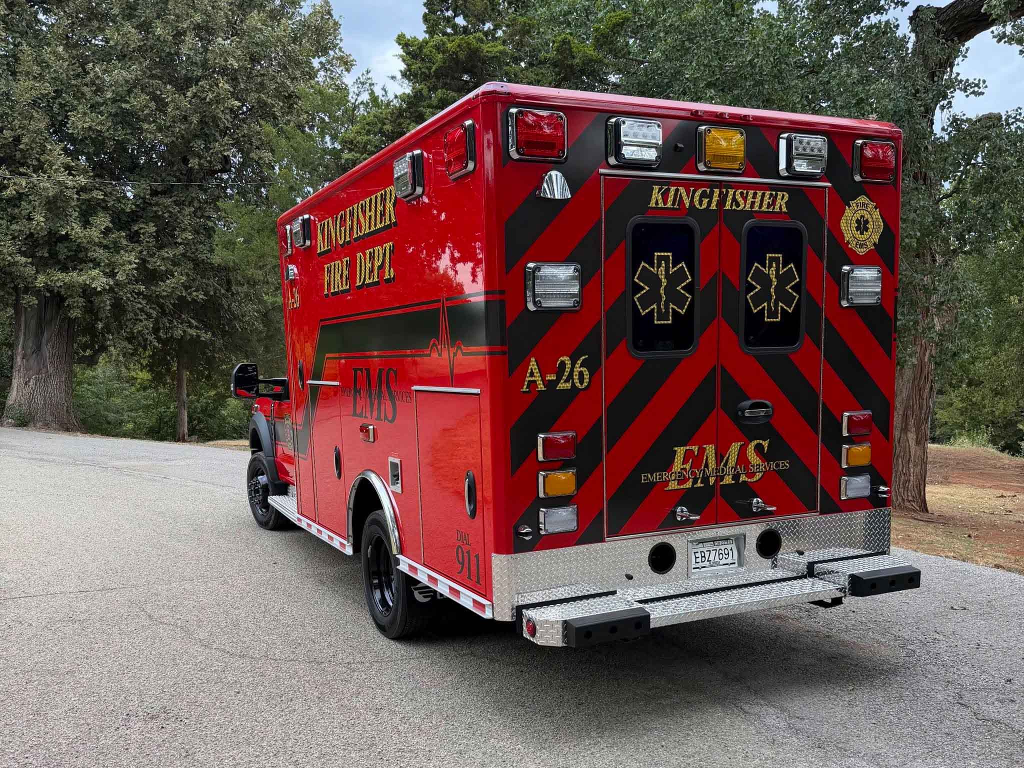Rear and side view of a white emergency ambulance with red and black chevron stripes and multiple roof lights, parked on a street.